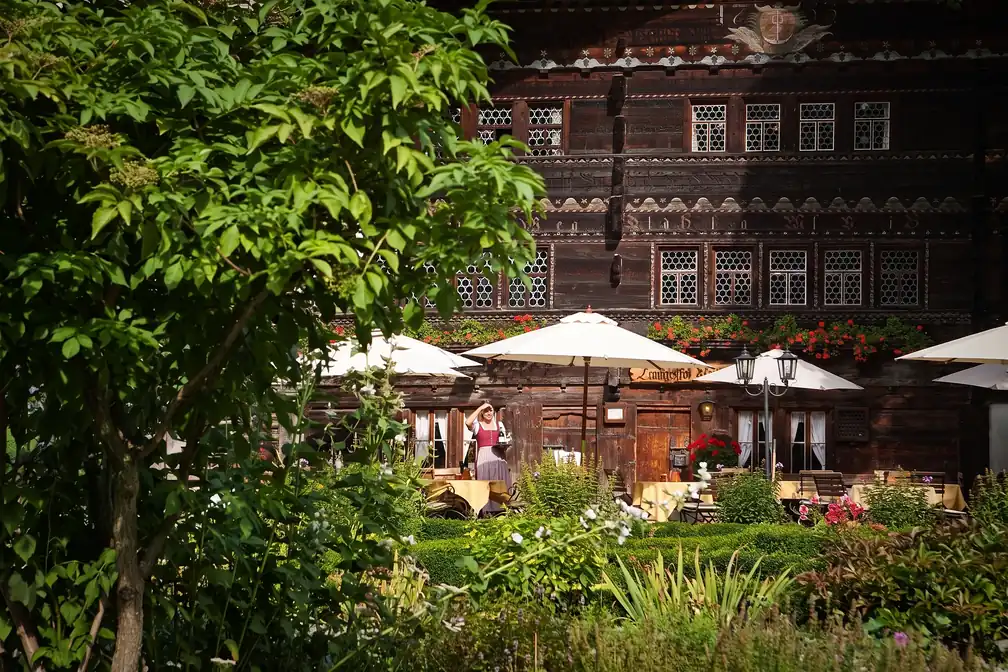 Fassade hinter Laub, davor die Terrasse mit Schirmen